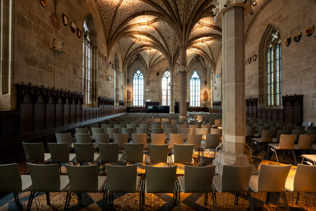 Historic hall inside Bebenhausen Abbey with Gothic vaulted ceilings, stained glass windows, ornate stone architecture, and seating area.