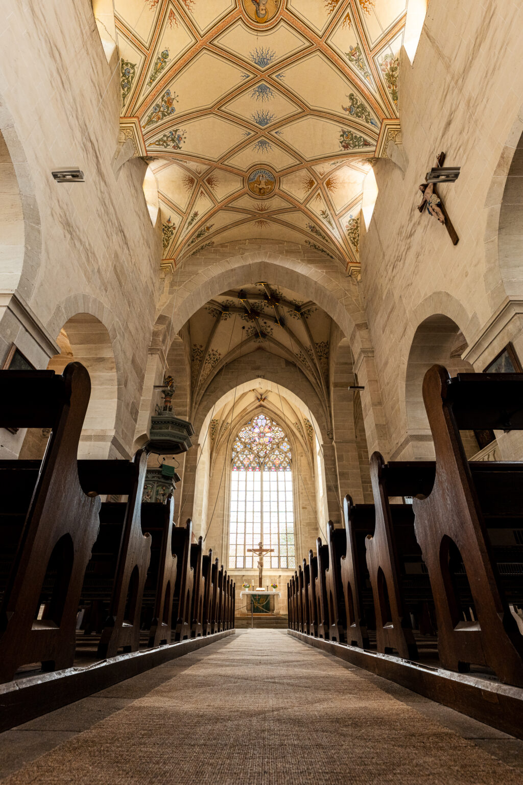Interior of Bebenhausen Abbey church with Gothic arches, stained glass window, wooden pews, and ornate vaulted ceiling.